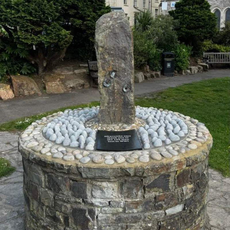 Image of the War Memorial at Runnymede Gardens in Ilfracombe. Image shows a stone circular base the top of which is inset with lightly coloured pebbles featuring a plaque and a vertical slab of stone which has 3 poppies embedded into it.